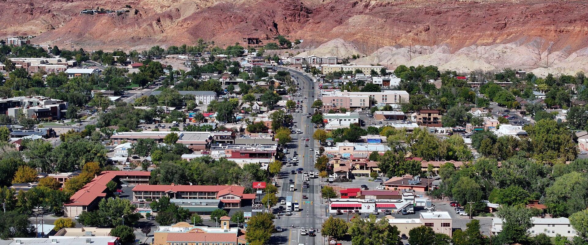 An elevated, wide-angle view of a town center street lined with low-rise commercial buildings, parking lots, and green trees, set against a backdrop of a large, layered red rock mesa.