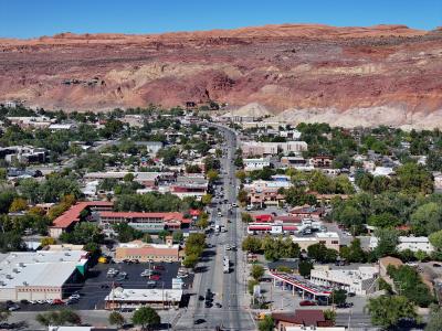 An elevated, wide-angle view of a town center street lined with low-rise commercial buildings, parking lots, and green trees, set against a backdrop of a large, layered red rock mesa.