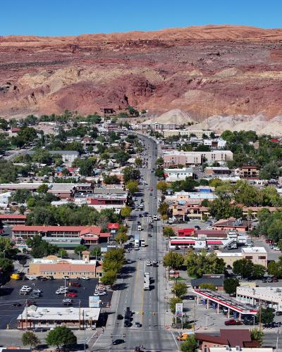 An elevated, wide-angle view of a town center street lined with low-rise commercial buildings, parking lots, and green trees, set against a backdrop of a large, layered red rock mesa.