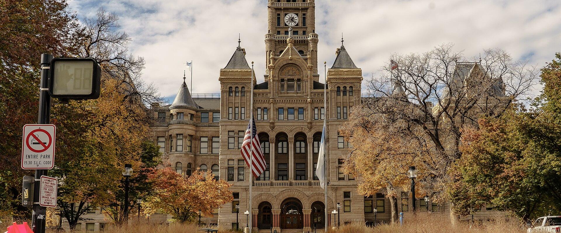 A large, ornate Romanesque Revival government building with a prominent central clock tower, viewed from a paved walkway lined with tall grasses and autumn trees.
