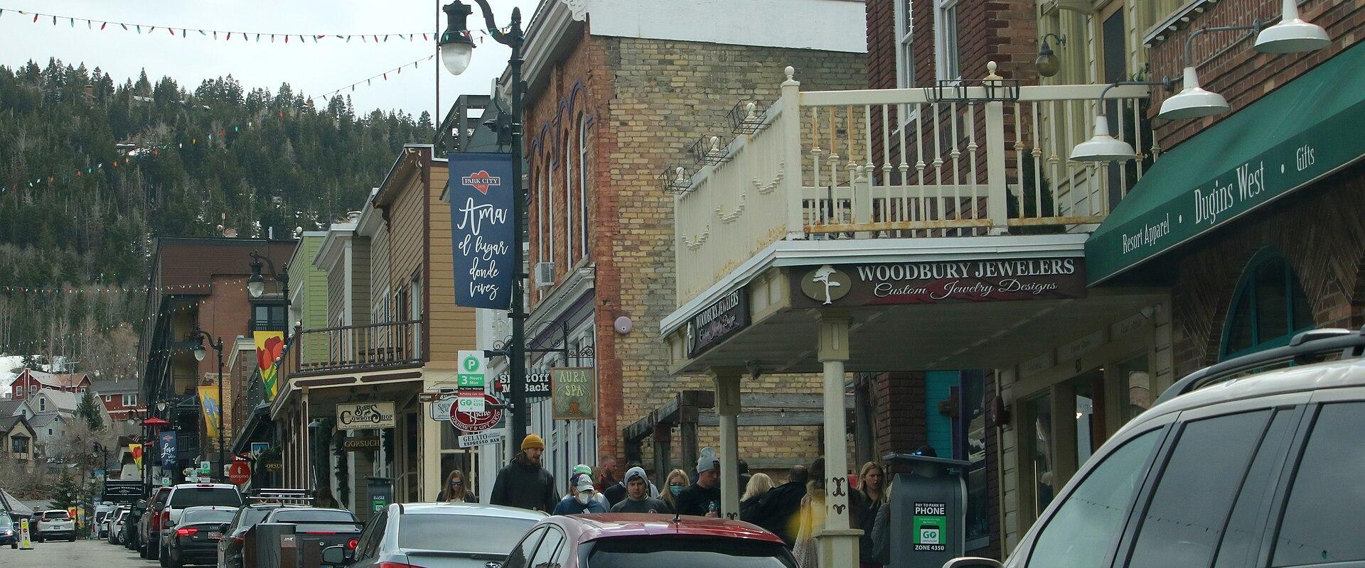 A street-level view of a commercial district with brick buildings, storefronts including "Woodburne Jewelers" and "D'Angelo's," parked cars, and a forested hill in the background.