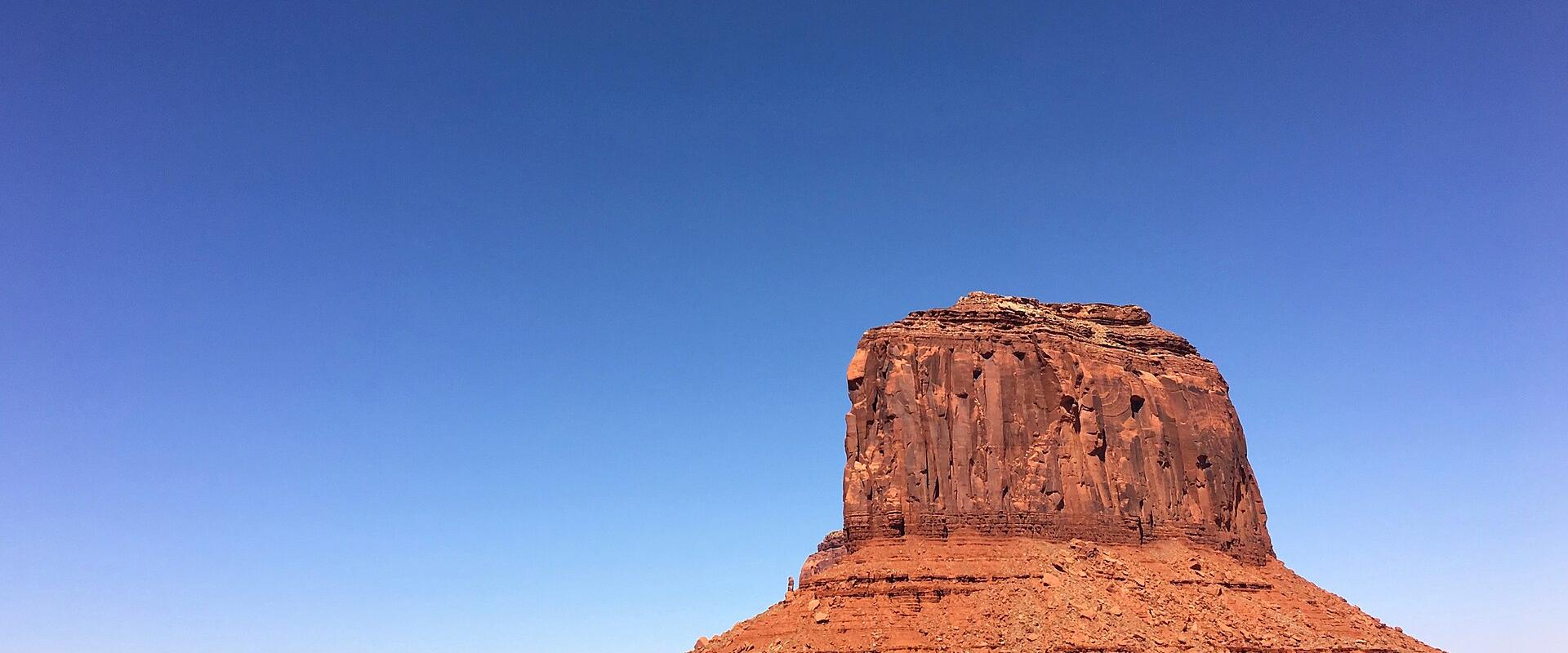 The image shows a large, isolated red sandstone butte (resembling the "Saddle" or "Saddle Rock" formation) rising from a flat, desert landscape with sparse vegetation, set against a clear blue sky.