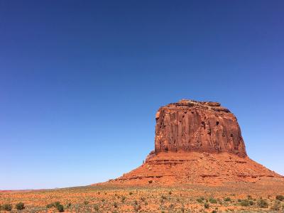 The image shows a large, isolated red sandstone butte (resembling the "Saddle" or "Saddle Rock" formation) rising from a flat, desert landscape with sparse vegetation, set against a clear blue sky.