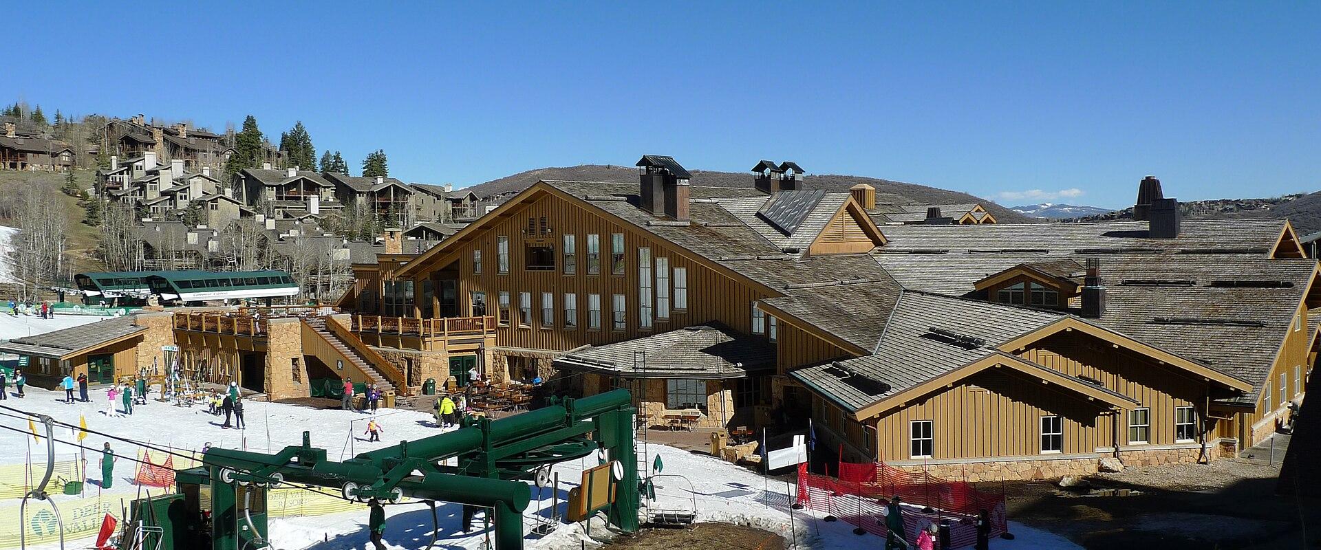 A view of a large, multi-story lodge with a wooden facade and a green chairlift in the foreground, set against a clear blue sky with snow on the ground.
