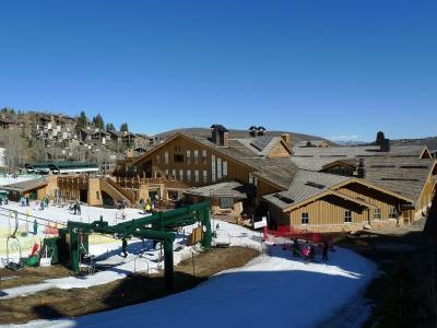 A view of a large, multi-story lodge with a wooden facade and a green chairlift in the foreground, set against a clear blue sky with snow on the ground.