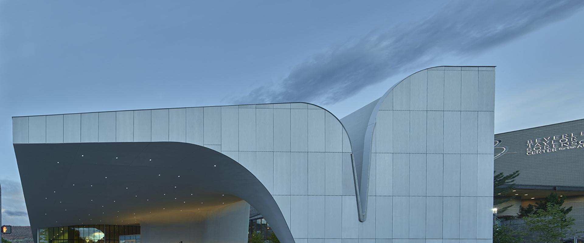 The image shows the exterior of a modern building with a white, undulating facade and a large overhanging canopy, with a few people standing near the entrance on a paved plaza.
