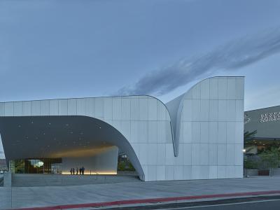 The image shows the exterior of a modern building with a white, undulating facade and a large overhanging canopy, with a few people standing near the entrance on a paved plaza.