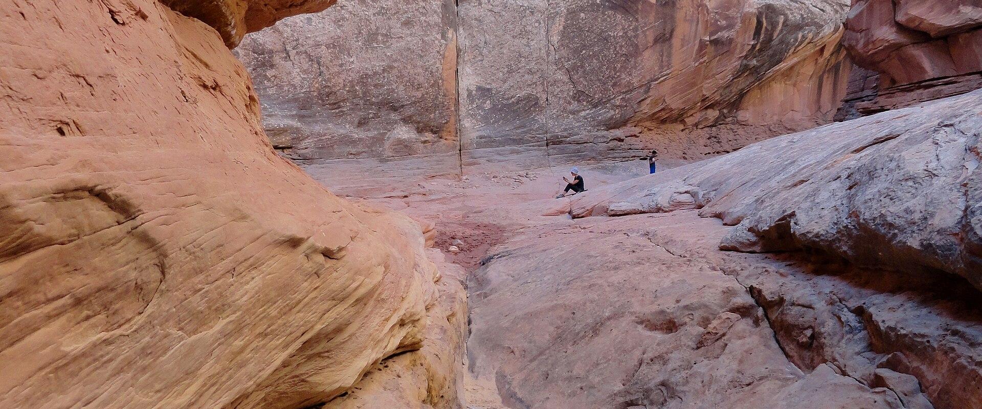 The image shows the interior of a narrow, winding sandstone slot canyon with smooth, sculpted walls, where two people are standing on the sandy floor in the distance.