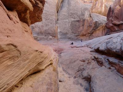 The image shows the interior of a narrow, winding sandstone slot canyon with smooth, sculpted walls, where two people are standing on the sandy floor in the distance.