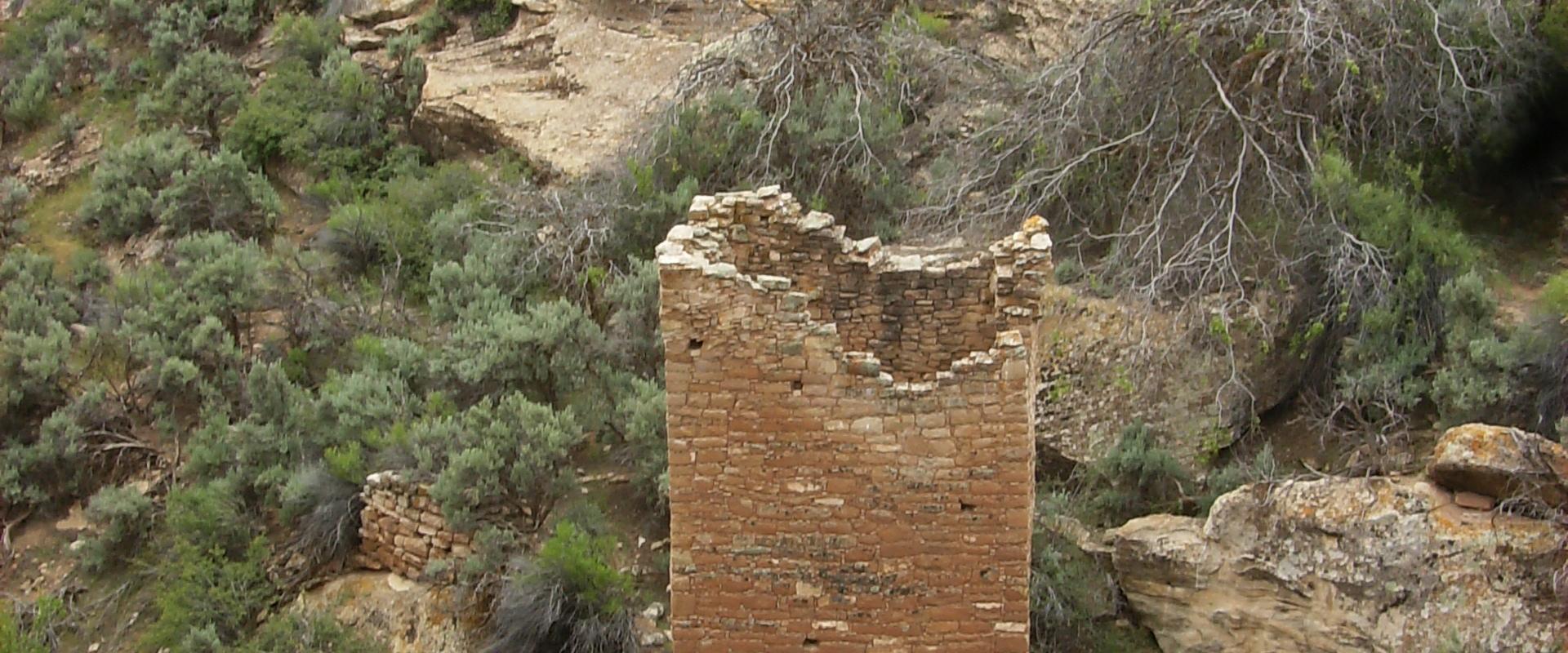 A stone tower structure (part of a larger ruin complex) situated in a desert landscape with green vegetation, viewed from a high vantage point.