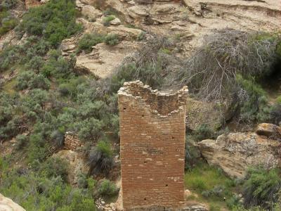 A stone tower structure (part of a larger ruin complex) situated in a desert landscape with green vegetation, viewed from a high vantage point.