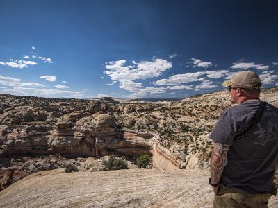 A man in a grey t-shirt and cap stands on a sandstone ledge overlooking a vast, arid canyon landscape with layered rock formations and sparse vegetation under a blue sky.