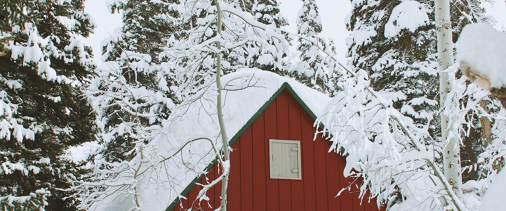 A red wooden building with a steep, snow-covered roof, surrounded by snow-laden evergreen trees and aspen branches in the foreground.