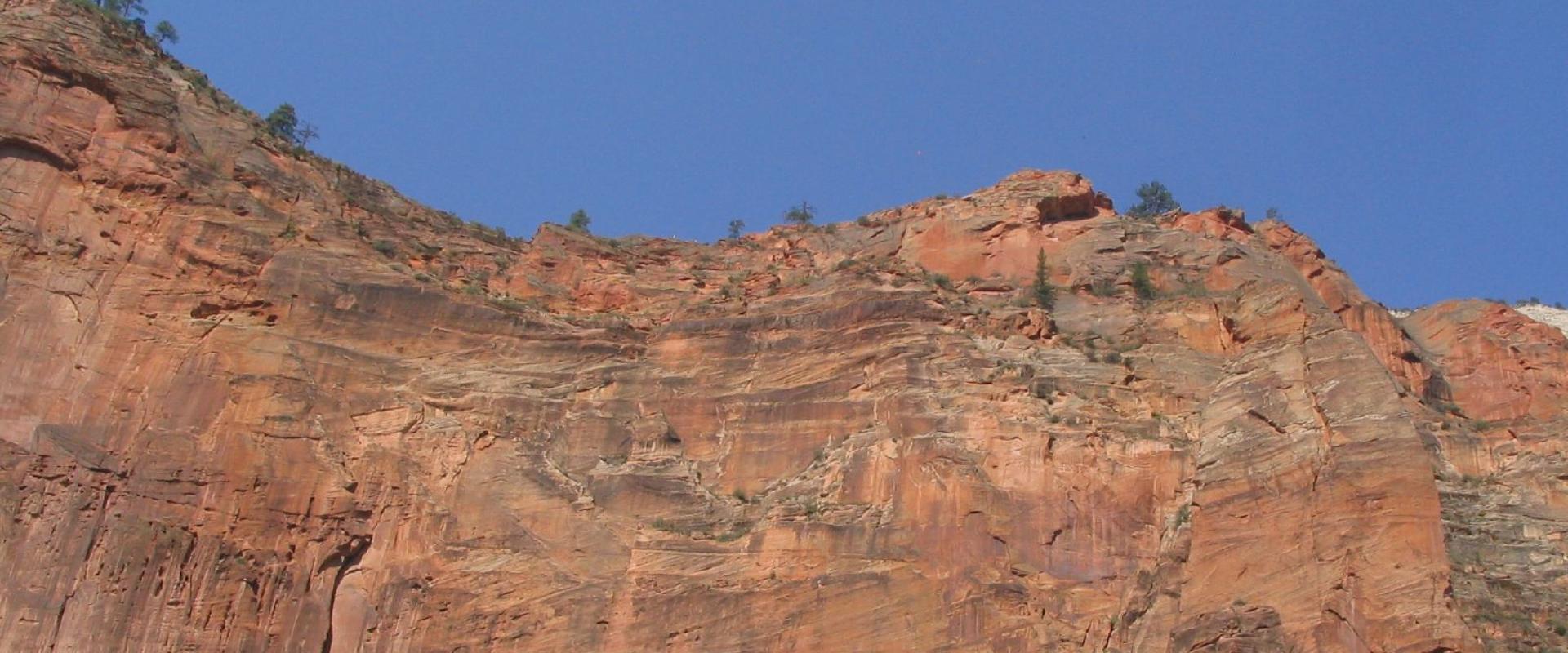 A low-angle view of a steep, reddish-orange sandstone cliff face with sparse vegetation, set against a clear blue sky.