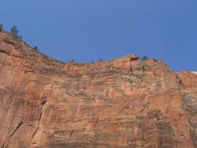 A low-angle view of a steep, reddish-orange sandstone cliff face with sparse vegetation, set against a clear blue sky.