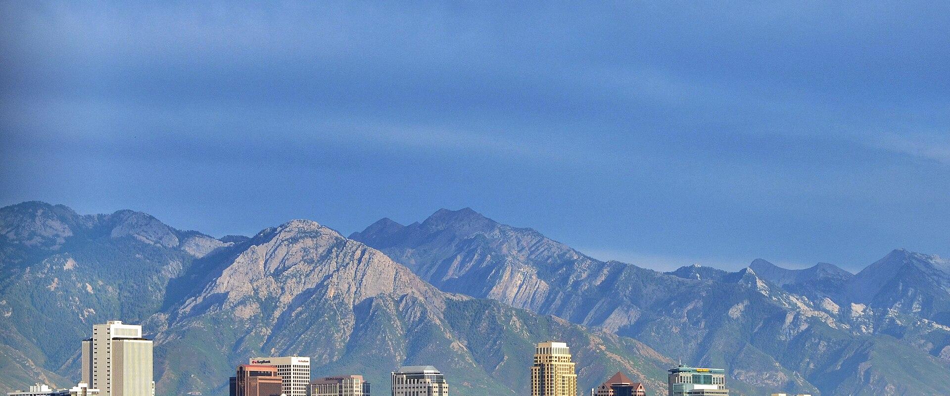 A city skyline with several high-rise buildings situated in front of a large, rugged mountain range under a blue sky.