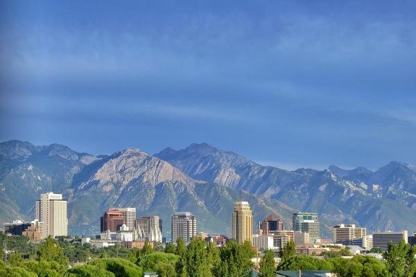 A city skyline with several high-rise buildings situated in front of a large, rugged mountain range under a blue sky.