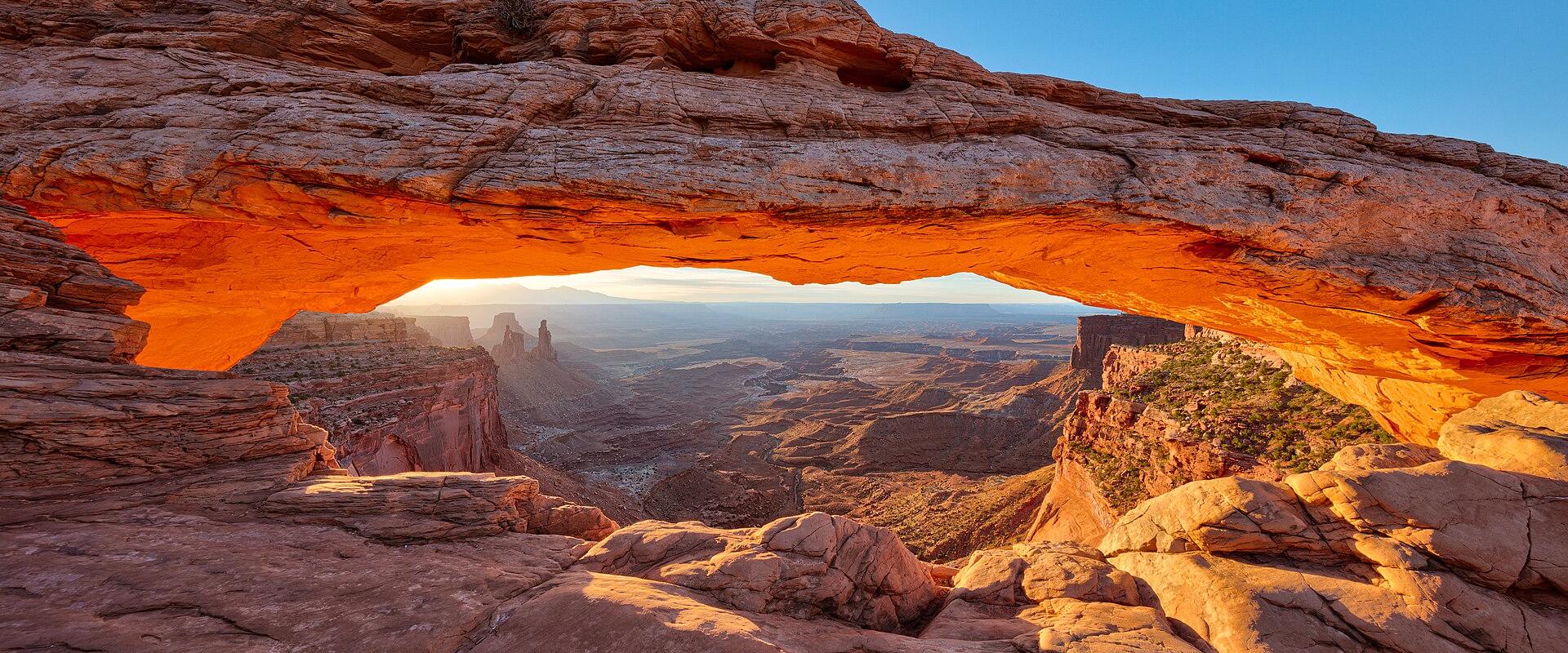 A low-angle view of a natural sandstone arch, with the sun rising or setting behind a distant butte, casting a warm orange glow on the rock face.