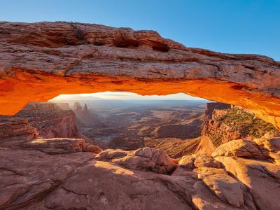 A low-angle view of a natural sandstone arch, with the sun rising or setting behind a distant butte, casting a warm orange glow on the rock face.