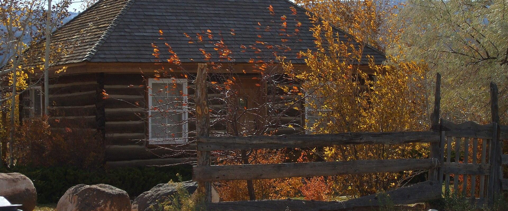A rustic, octagonal log cabin structure with a dark, multi-sided roof, situated behind a wooden split-rail fence, surrounded by trees with autumn foliage.