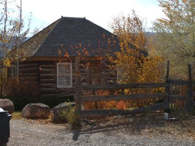 A rustic, octagonal log cabin structure with a dark, multi-sided roof, situated behind a wooden split-rail fence, surrounded by trees with autumn foliage.