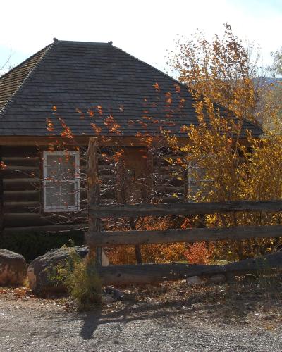 A rustic, octagonal log cabin structure with a dark, multi-sided roof, situated behind a wooden split-rail fence, surrounded by trees with autumn foliage.