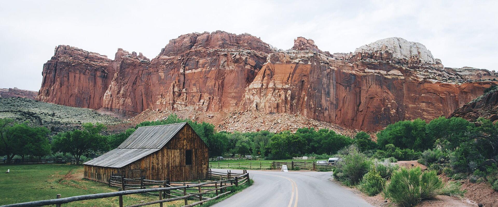A paved road curves to the right in the foreground, flanked by a wooden fence and green vegetation, leading towards a large, reddish-brown rock formation (the Hickory Bridge) in the background.