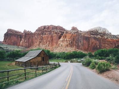 A paved road curves to the right in the foreground, flanked by a wooden fence and green vegetation, leading towards a large, reddish-brown rock formation (the Hickory Bridge) in the background.