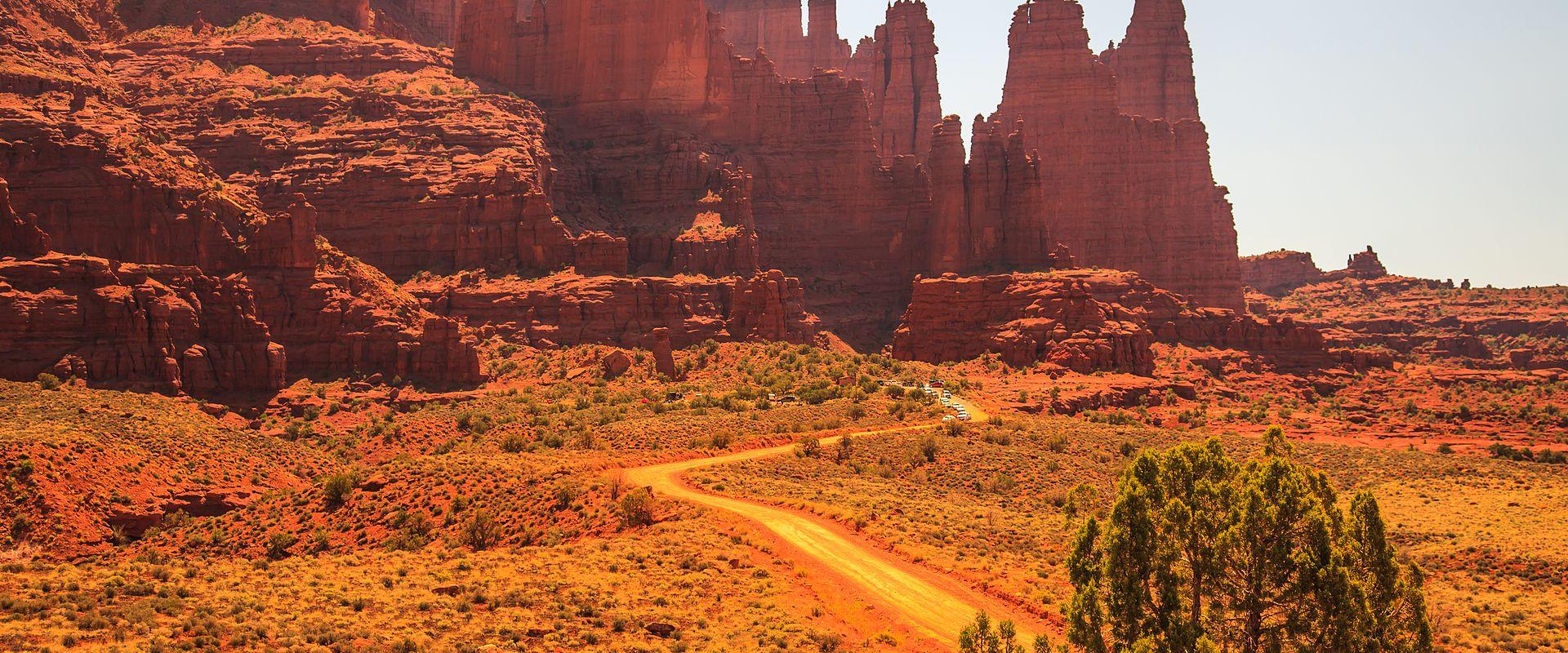 A landscape photograph showing a winding dirt road leading through a desert valley towards a cluster of massive, vertical red sandstone spires and cliffs under a clear sky.