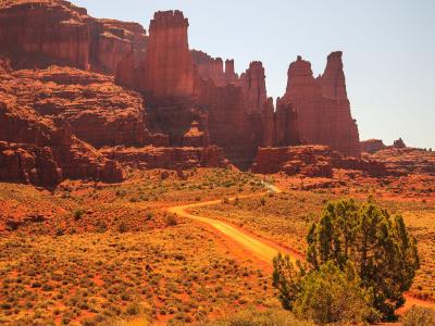 A landscape photograph showing a winding dirt road leading through a desert valley towards a cluster of massive, vertical red sandstone spires and cliffs under a clear sky.