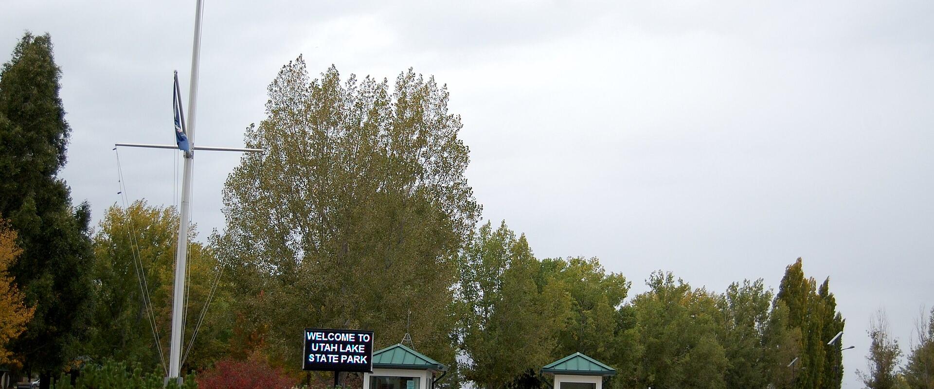 The image shows the entrance to a park with a flagpole, two guard booths, and a black sign reading "WELCOME TO UTAH LAKE STATE PARK".