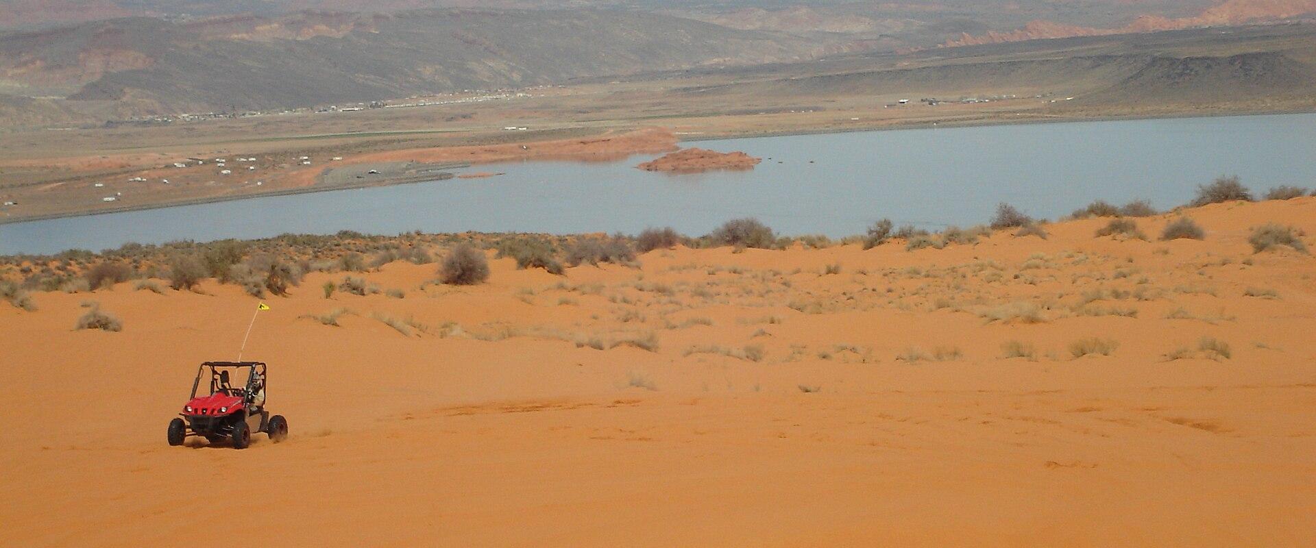 A wide landscape view showing a red dune buggy driving on orange sand dunes in the foreground, with a large body of water (a reservoir) and distant mountains in the background.