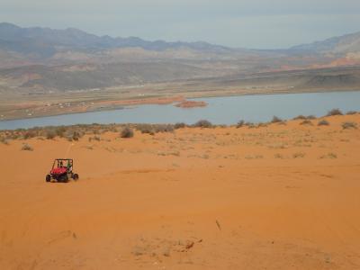 A wide landscape view showing a red dune buggy driving on orange sand dunes in the foreground, with a large body of water (a reservoir) and distant mountains in the background.