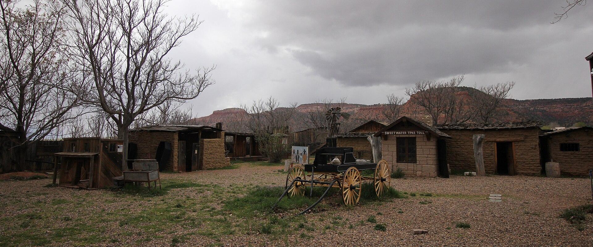 An outdoor scene showing a collection of rustic, adobe-style buildings and a black stagecoach with large wooden wheels, set against a backdrop of red rock cliffs and a cloudy sky.
