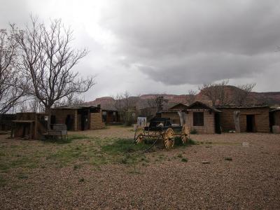 An outdoor scene showing a collection of rustic, adobe-style buildings and a black stagecoach with large wooden wheels, set against a backdrop of red rock cliffs and a cloudy sky.