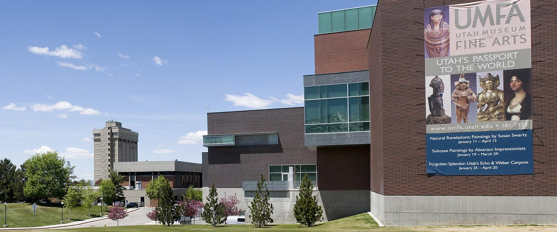 The image shows the exterior of a modern brick and glass building with a large banner on the facade reading "UMFA UTAH MUSEUM OF FINE ARTS" and listing current exhibitions.