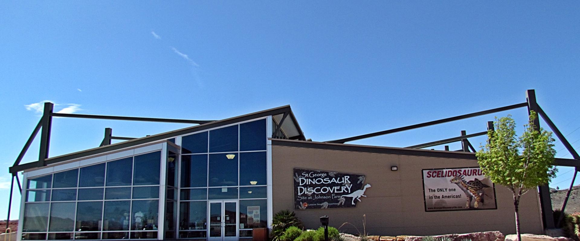 The image shows the exterior of a modern building with a large glass facade and a flat roof, featuring a prominent sign that reads "St. George Dinosaur Discovery Site" along with a graphic of a dinosaur skeleton.