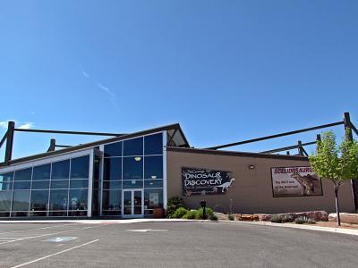The image shows the exterior of a modern building with a large glass facade and a flat roof, featuring a prominent sign that reads "St. George Dinosaur Discovery Site" along with a graphic of a dinosaur skeleton.