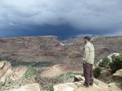 A man in a cap and light jacket stands on a rocky outcrop in the foreground, looking out over a deep, wide canyon with layered red rock walls and a river visible in the distance, under a dramatic, cloudy sky.