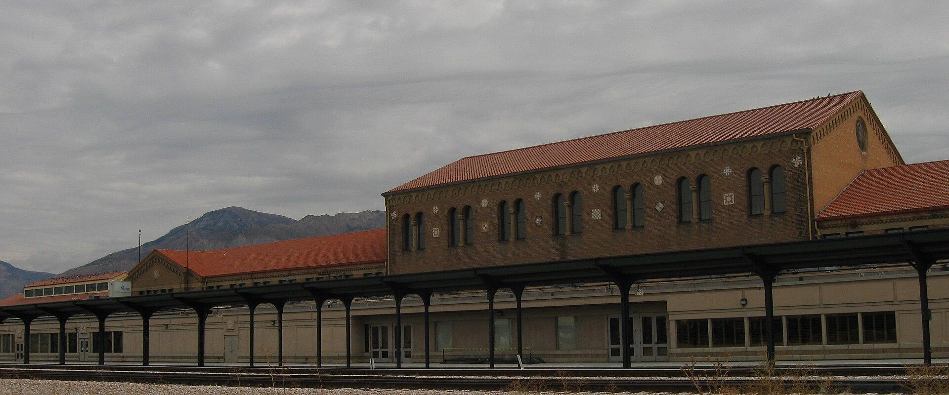 The image shows a long, low, brick building with a terracotta-tiled hipped roof, situated next to railroad tracks, with a mountain range visible in the background.