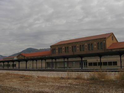 The image shows a long, low, brick building with a terracotta-tiled hipped roof, situated next to railroad tracks, with a mountain range visible in the background.
