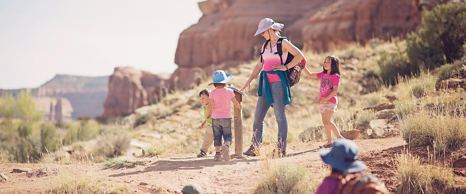 A group of adults and children hiking on a dirt trail in a red rock desert landscape, with one person interacting with a wooden post or signpost.