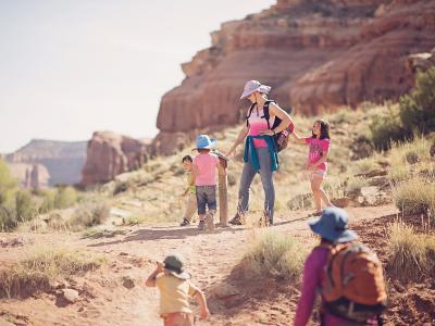 A group of adults and children hiking on a dirt trail in a red rock desert landscape, with one person interacting with a wooden post or signpost.