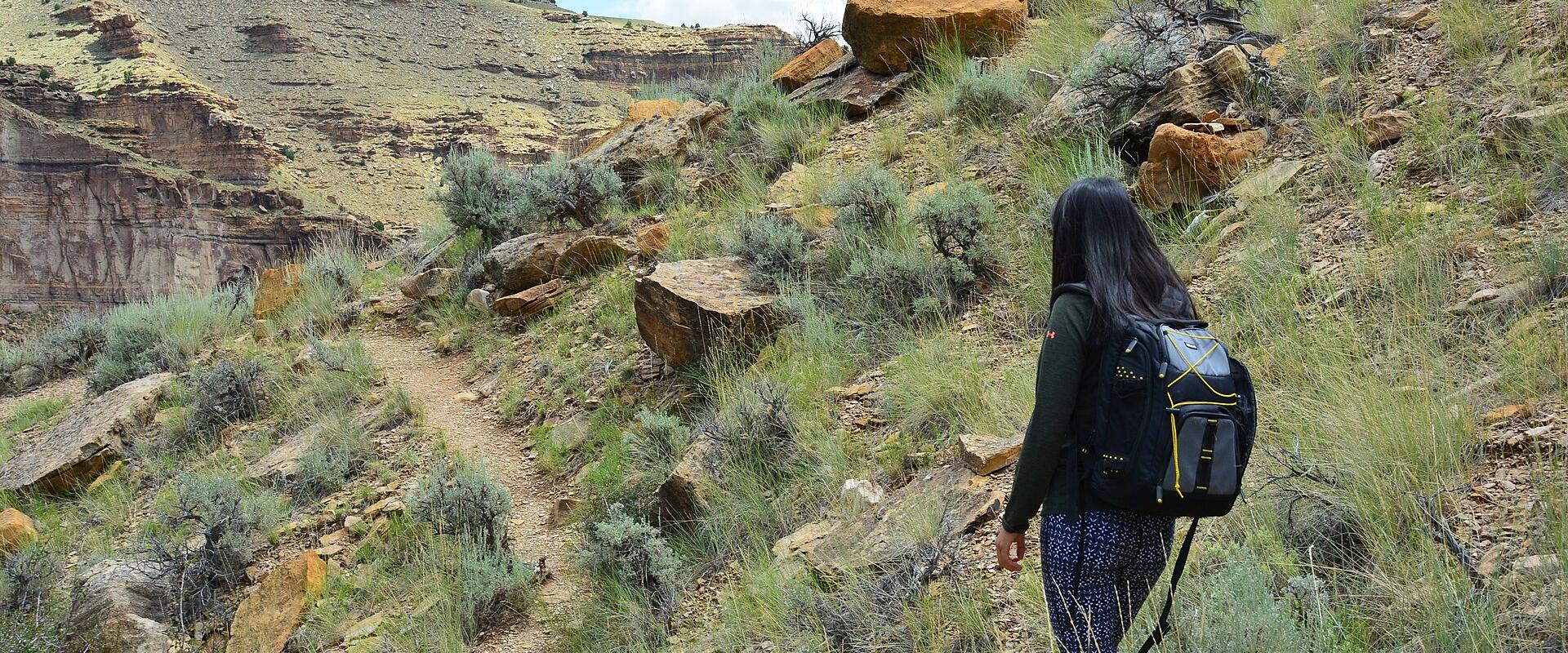 A hiker with a backpack walks along a dirt trail on a grassy, rocky slope overlooking a canyon with layered rock formations.
