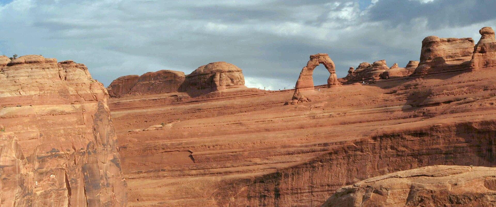 The image shows a large, freestanding natural sandstone arch (Delicate Arch) set against a blue sky with clouds, situated within a landscape of red rock formations and desert terrain.