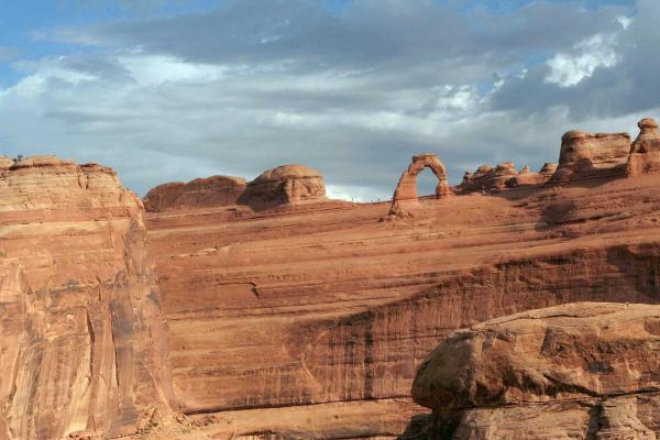 The image shows a large, freestanding natural sandstone arch (Delicate Arch) set against a blue sky with clouds, situated within a landscape of red rock formations and desert terrain.