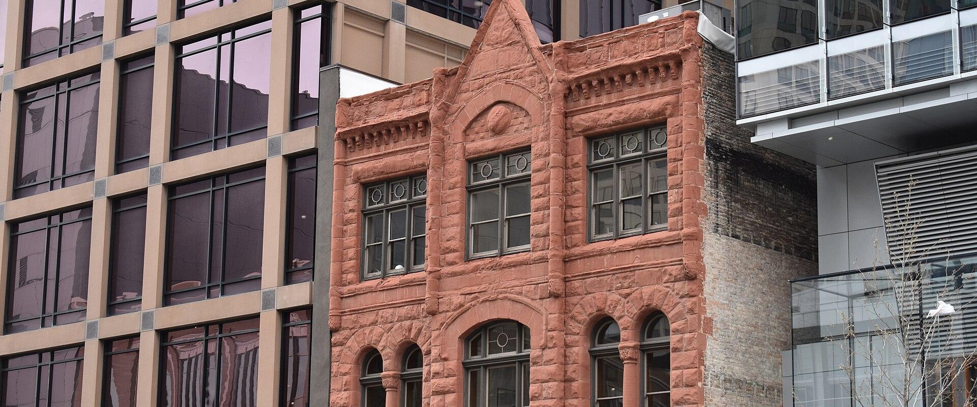 A narrow, three-story red sandstone building with distinctive Romanesque Revival architecture, featuring arched windows on the lower level and rectangular windows above, situated between two modern glass-and-steel office buildings; a partial sign rea