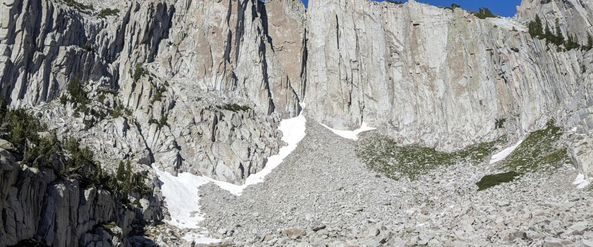 A high-angle view of a rocky cirque with steep granite walls, patches of snow, and a scree slope under a clear blue sky.