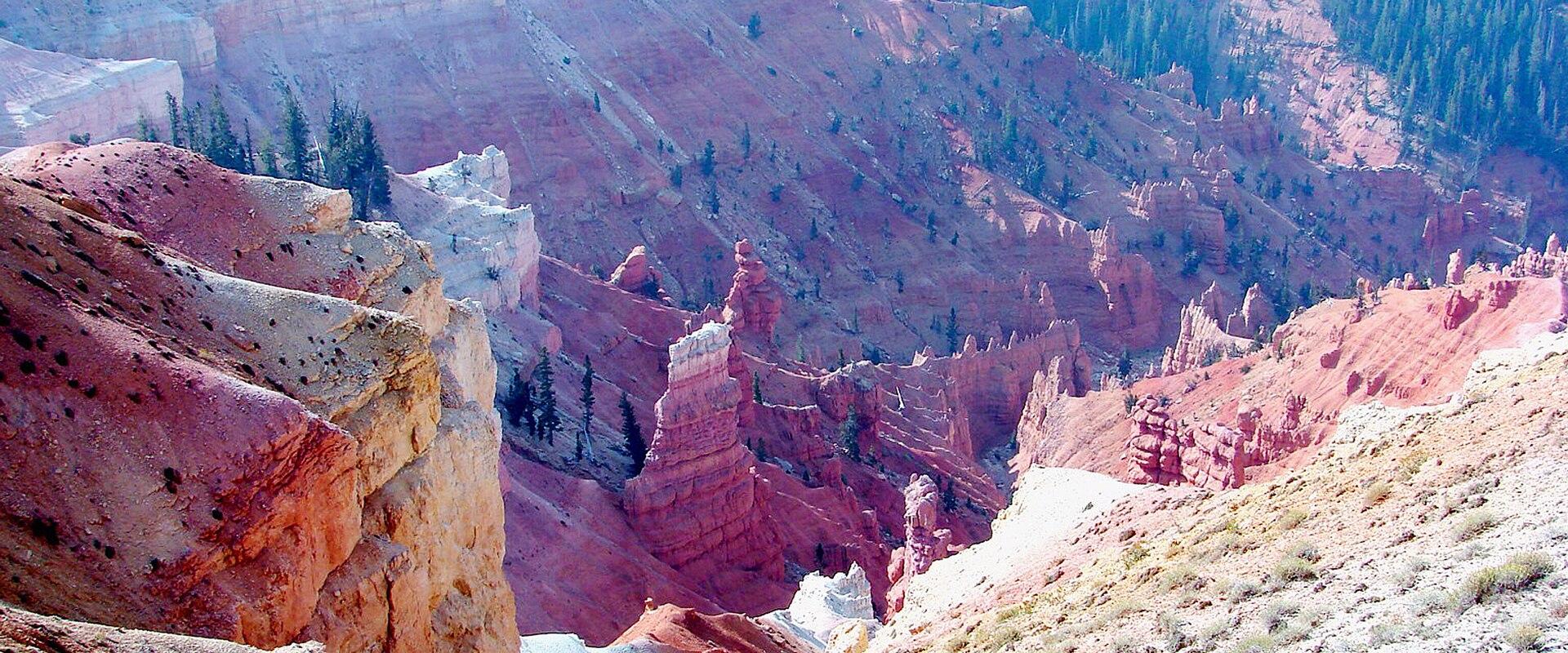 A high-angle view of a deep, amphitheater-style canyon with steep, multi-colored rock walls (red, white, and tan) and sparse vegetation, including a prominent hoodoo in the center, with a patch of snow visible in the lower center.
