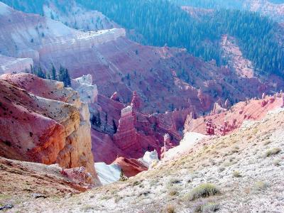 A high-angle view of a deep, amphitheater-style canyon with steep, multi-colored rock walls (red, white, and tan) and sparse vegetation, including a prominent hoodoo in the center, with a patch of snow visible in the lower center.